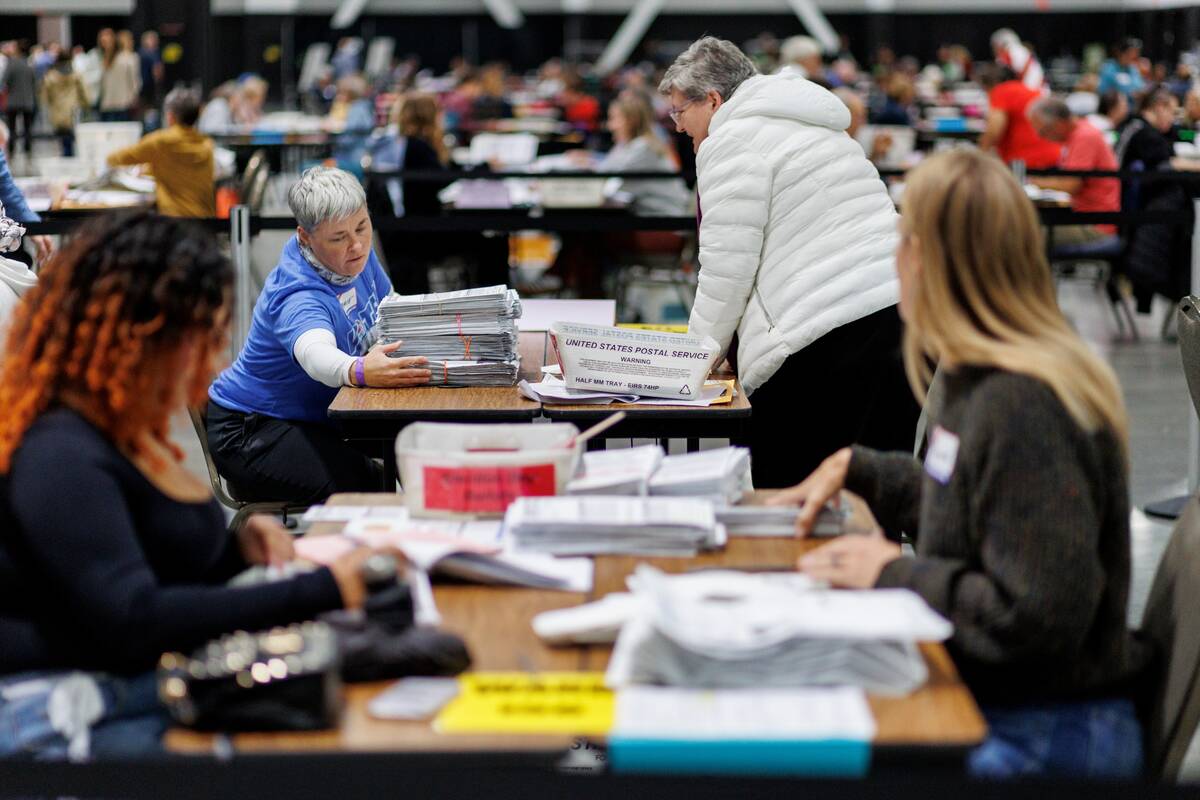 A group of adults work around a table in a large warehouse with paper ballots.